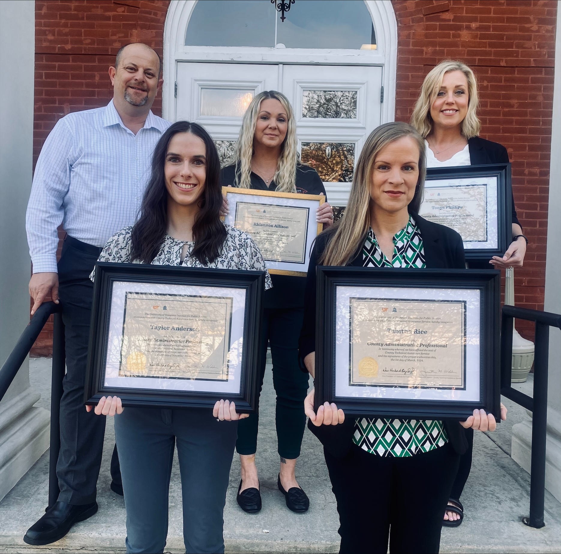 The image shows a group of five individuals standing on steps in front of a building with a red brick facade and white-framed windows. Four are holding a framed certificate indicating their completion of the County Administrative Professional program.